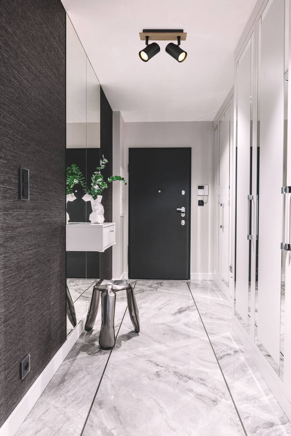 Entrée moderne avec plafonnier double spot noir éclairant le couloir, porte noire et sol marbre.