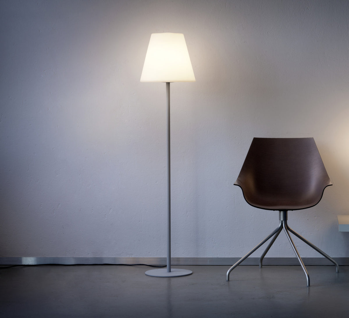 Lampadaire sur pied allumé en intérieur, abat-jour conique, mur gris et chaise brune