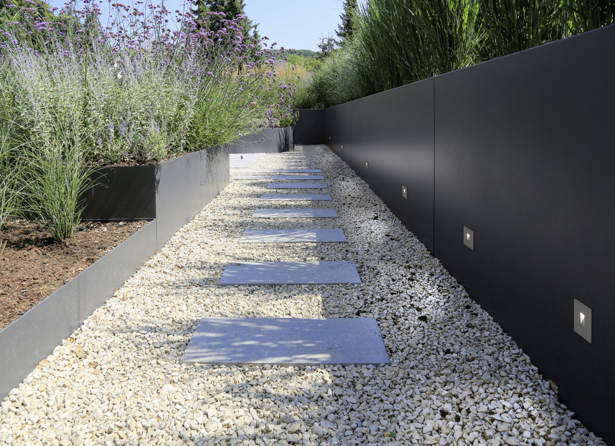 Cheminement de graviers blancs longeant un mur sombre équipé de plusieurs appliques