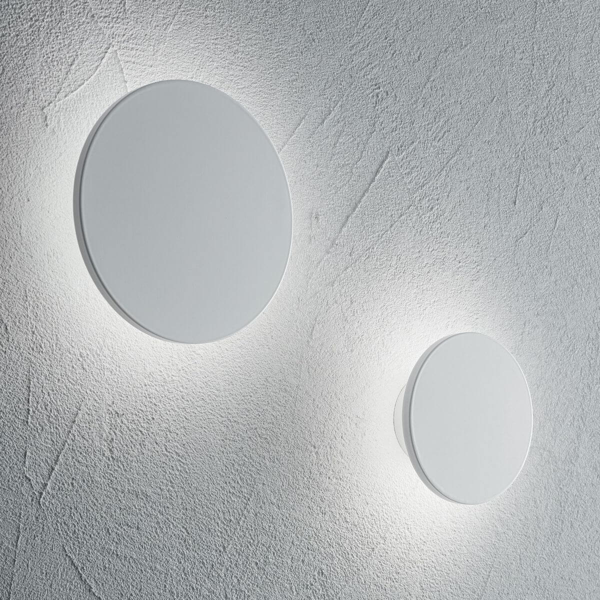 Deux appliques rondes blanches allumées sur un mur gris texturé, lumière diffuse