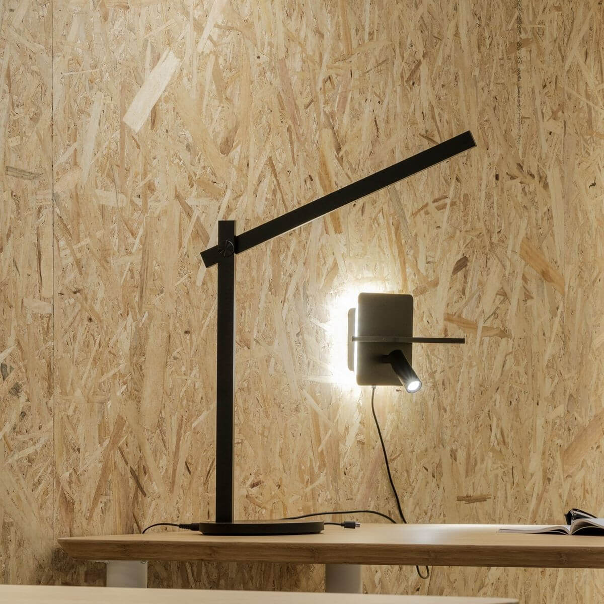Lampe noire en ambiance sur bureau en bois, éclairant un objet mural blanc.