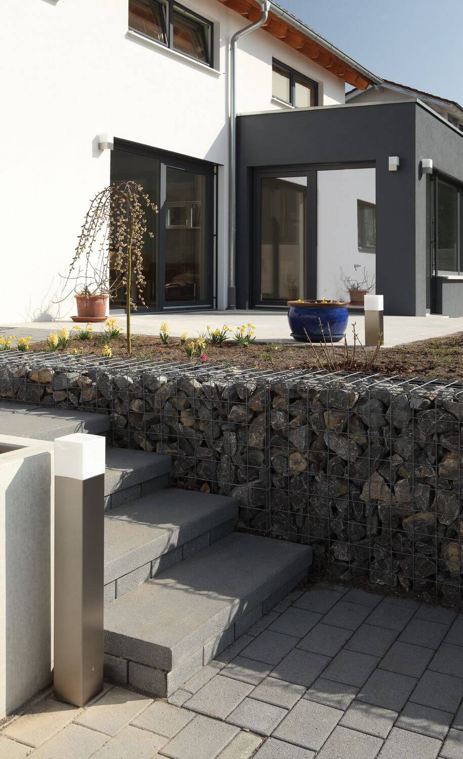 Deux bornes extérieures en inox éclairent une terrasse avec gabion et maison contemporaine