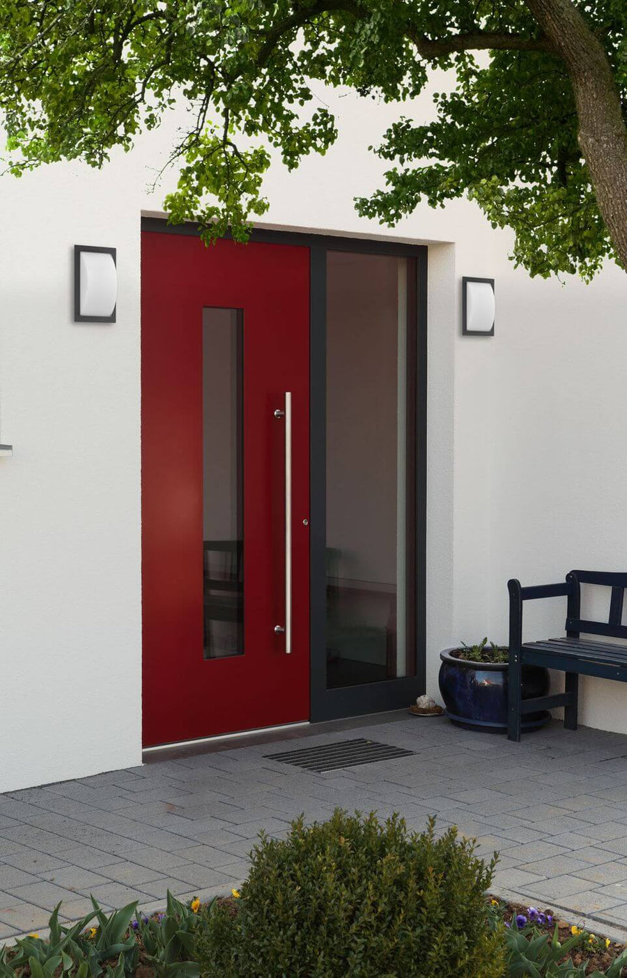Entrée moderne avec porte rouge, appliques anthracite et banc sous un arbre.