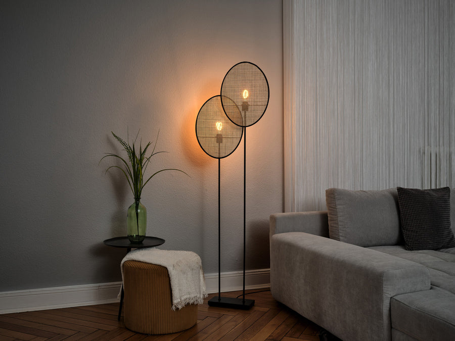 Lampadaire allumé dans un salon, près d’un canapé gris et d’une plante.