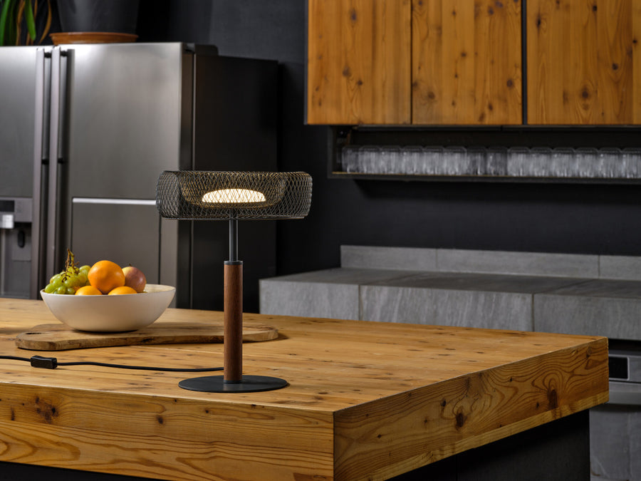 Lampe allumée sur îlot de cuisine en bois, bol de fruits et réfrigérateur en arrière-plan.