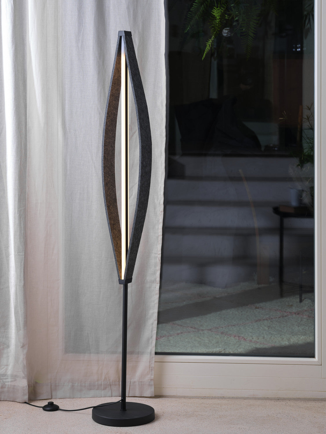 Lampadaire allumé près d’un rideau clair, devant une baie vitrée reflétant un intérieur sombre.