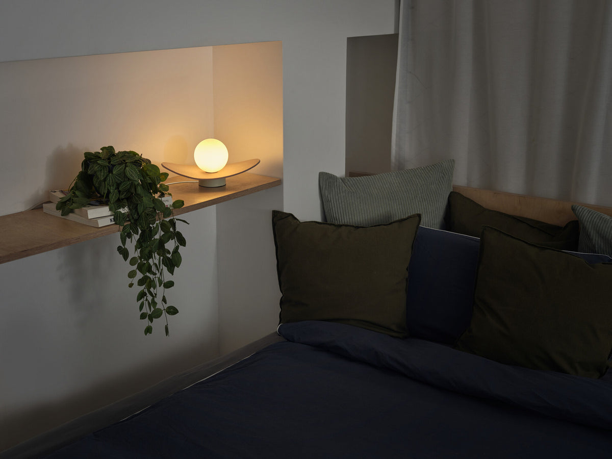 Lampe allumée sur étagère en bois près d'une plante et de coussins, ambiance chambre.