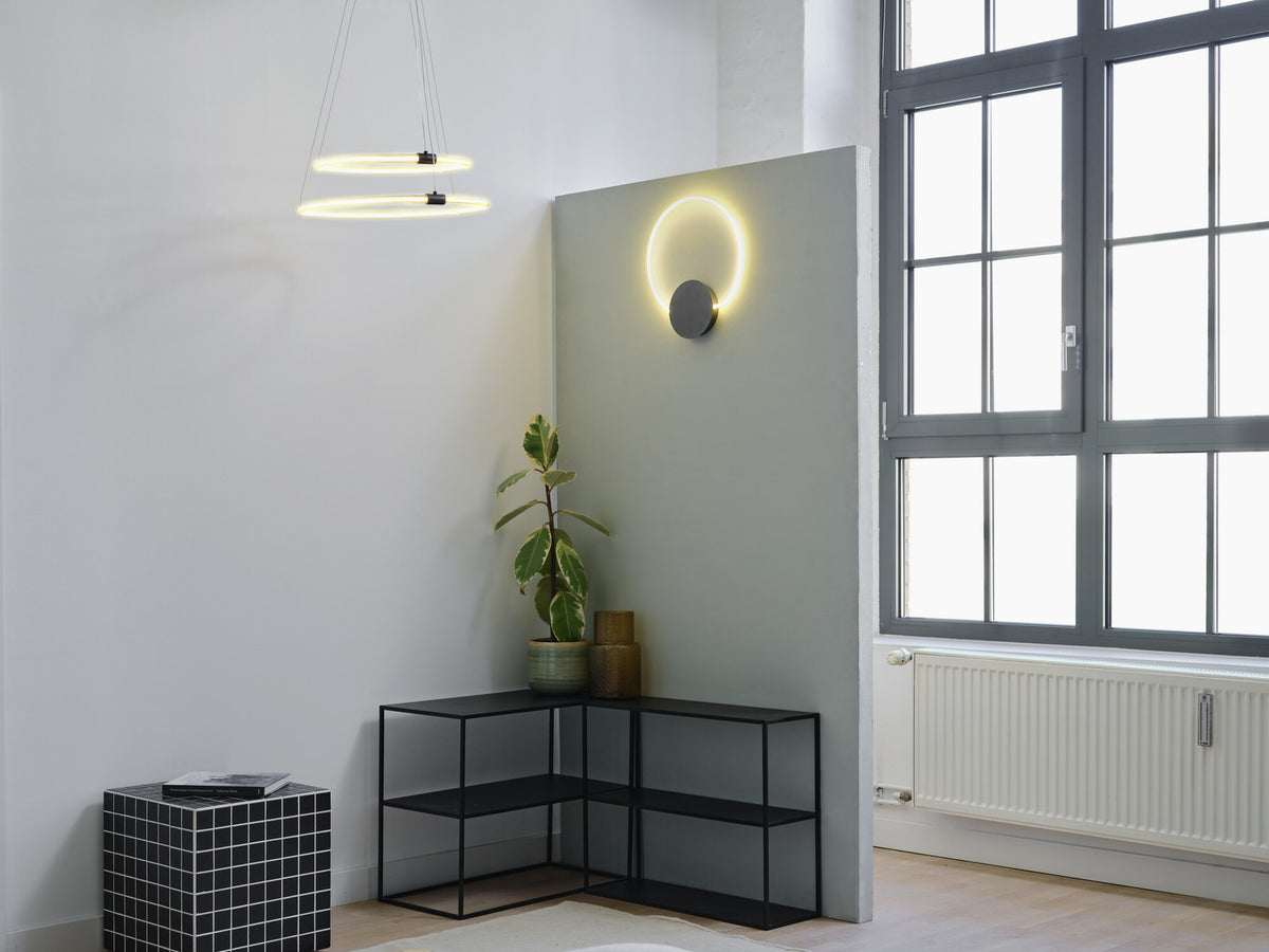 Mise en ambiance : applique ronde lumineuse sur cloison grise, suspension annulaire et étagère noire.