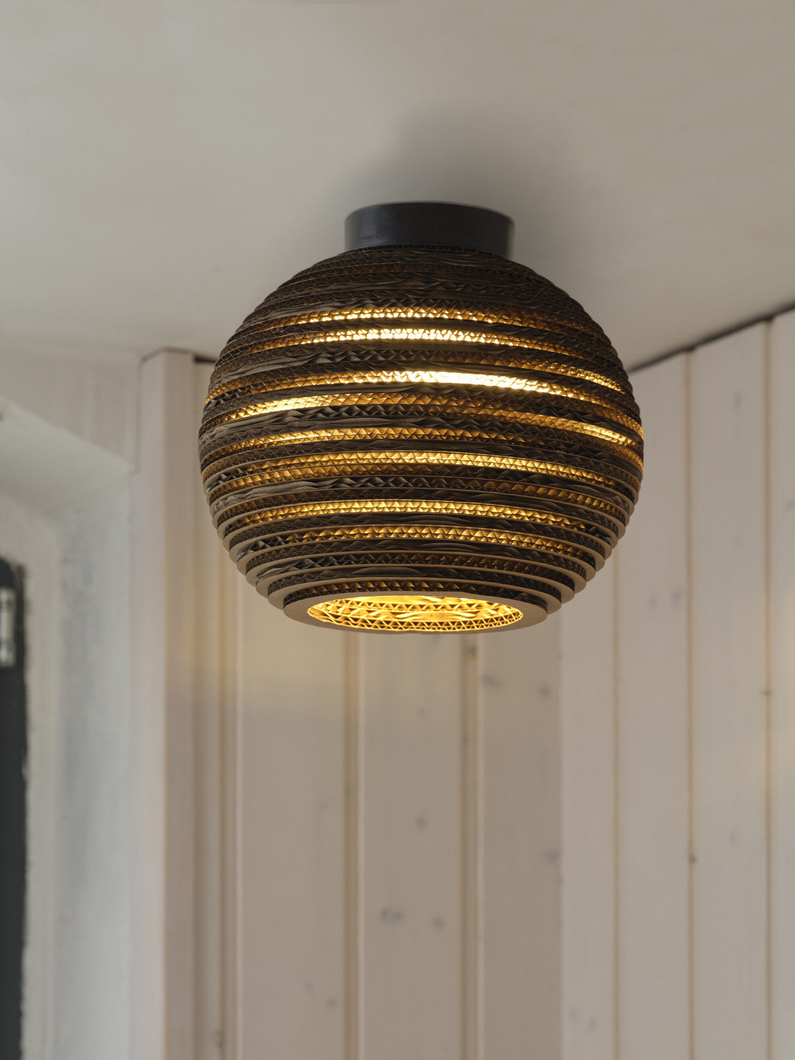 Détail du plafonnier allumé, lumière chaude filtrant entre les lamelles de carton.