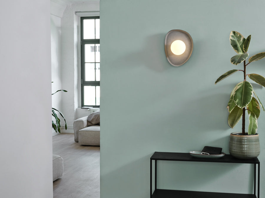 Applique murale allumée sur un mur vert, près d’une plante et d’une console.