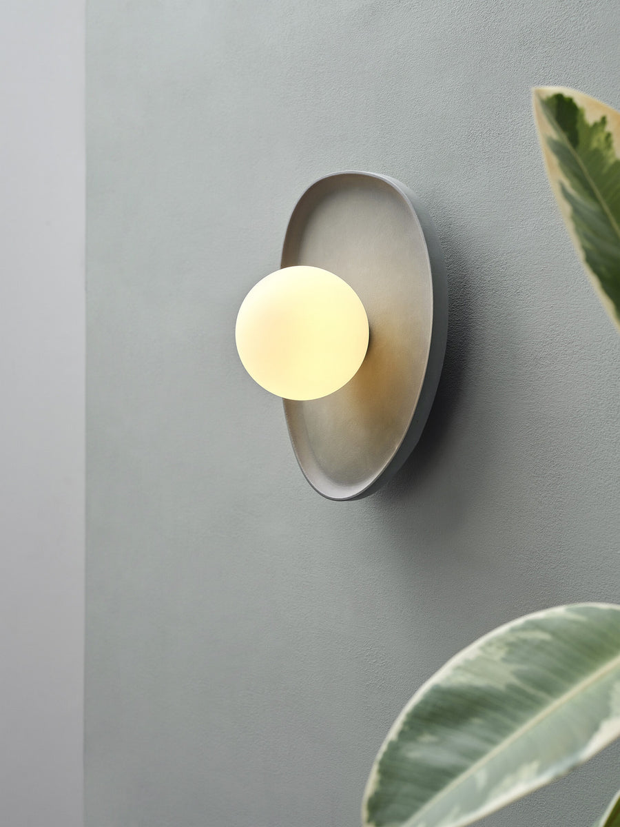 Gros plan de l’applique allumée sur un mur gris, avec feuillage flou.
