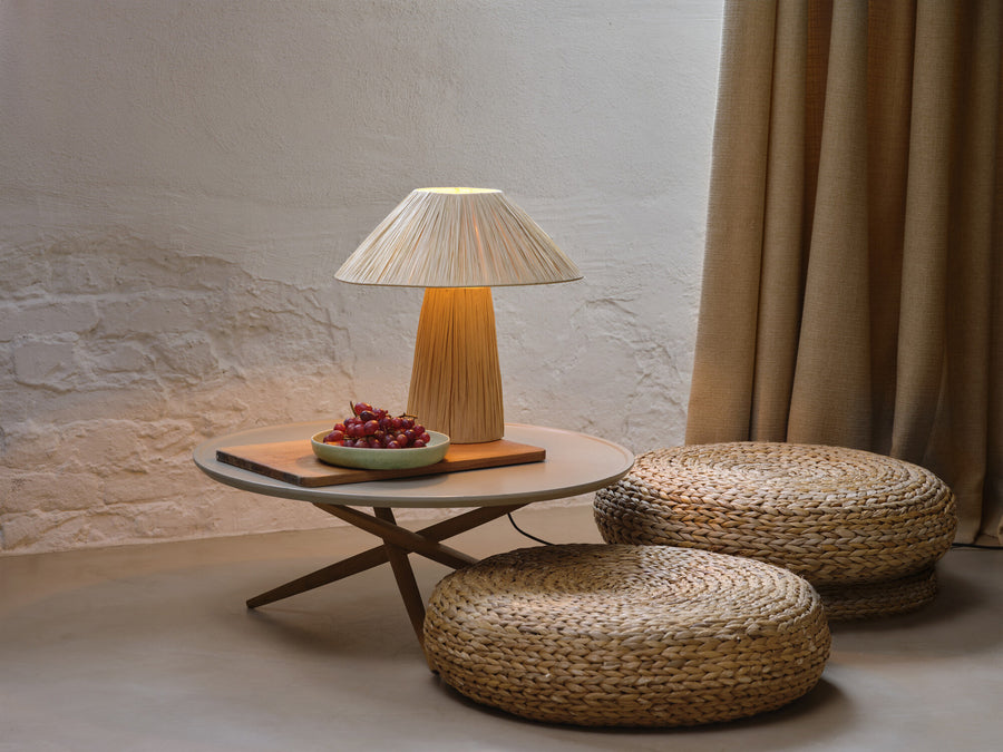 Lampe à poser allumée sur table basse, corbeille de raisins et poufs en sisal.