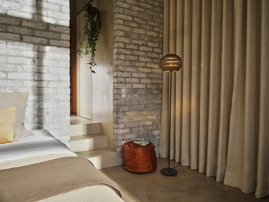 Lampadaire allumé dans une chambre, près d’un mur en briques et d’un pouf.