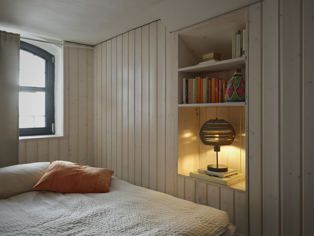 Lampe allumée dans une niche en bois clair, posée sur des livres près d’un lit.
