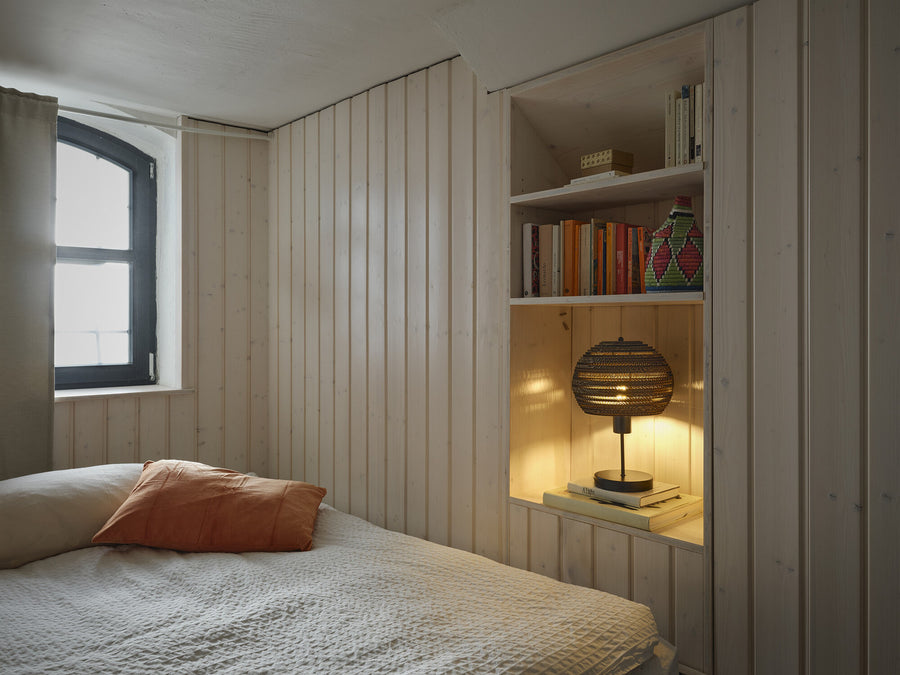 Lampe allumée dans une niche en bois clair, posée sur des livres près d’un lit.