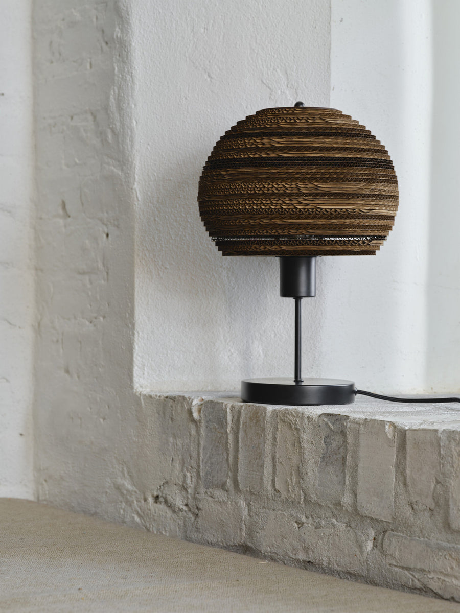 Lampe à poser près d’un mur blanc, pied noir et abat-jour sphérique en carton brun.