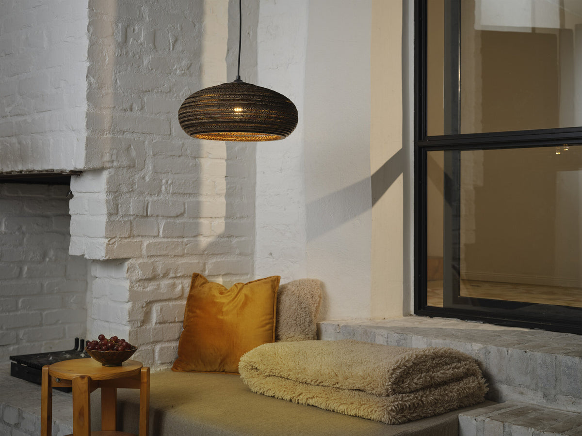 Mise en ambiance salon, suspension allumée devant un mur de briques blanches, coussins moutarde