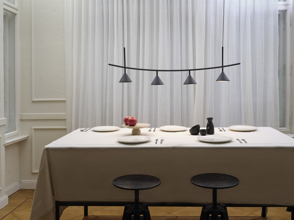 Mise en ambiance au-dessus d’une table dressée, suspension noire à quatre cônes devant des rideaux clairs.