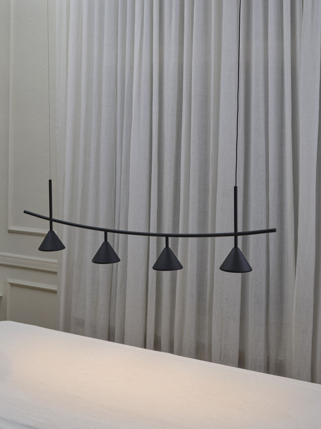 Détail de la suspension noire et de ses cônes devant des rideaux et une table claire.
