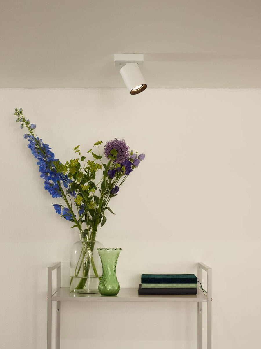 Plafonnier blanc orientable éclairant un mur avec vase de fleurs