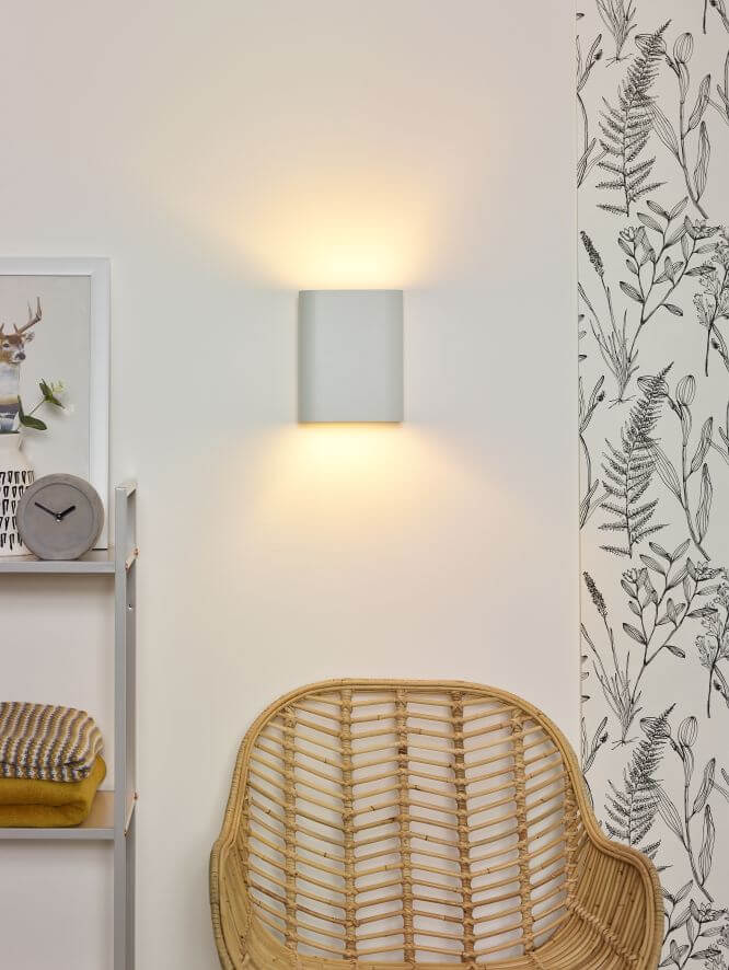 Applique murale allumée dans un salon, fauteuil rotin et papier peint végétal.