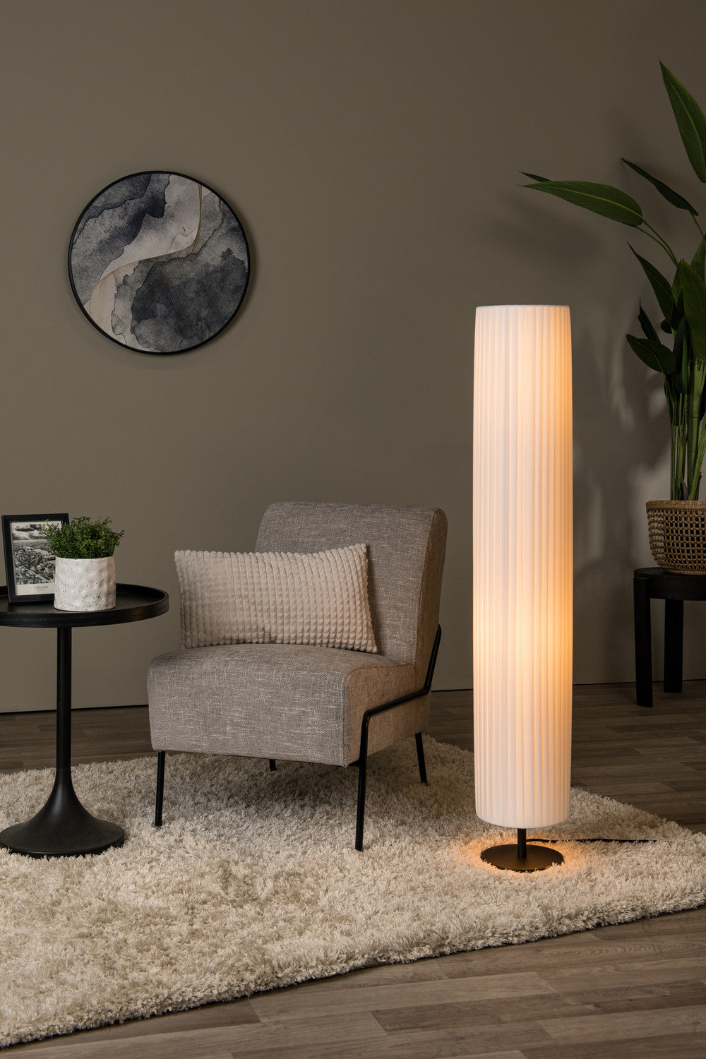 Mise en ambiance salon avec lampadaire cylindrique blanc allumé contre un mur taupe