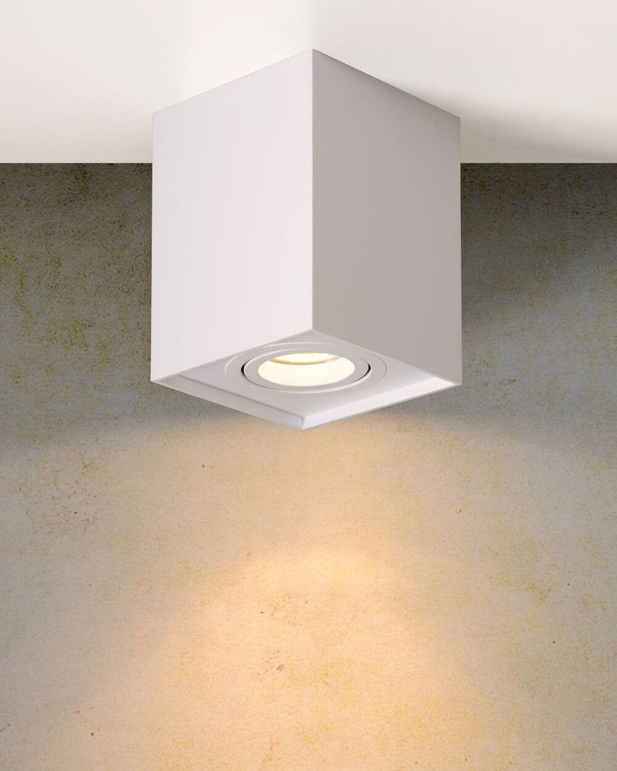 Plafonnier cube blanc allumé, projetant une lumière chaude sur un mur texturé beige.