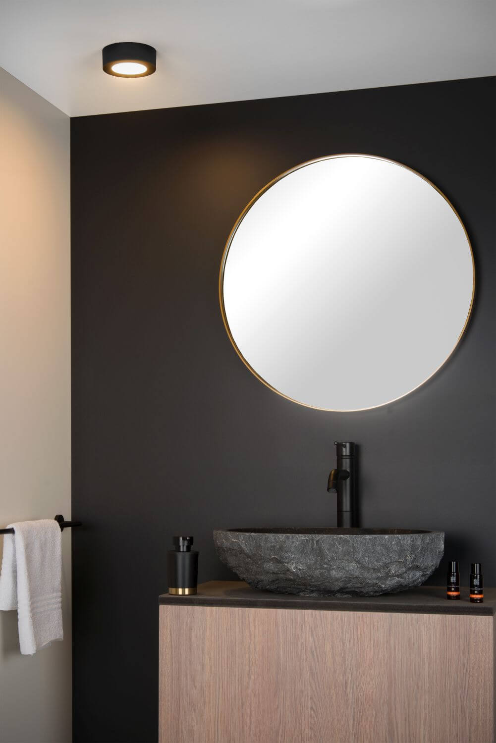 Plan rapproché du plafonnier noir allumé au plafond, miroir rond et vasque en pierre.