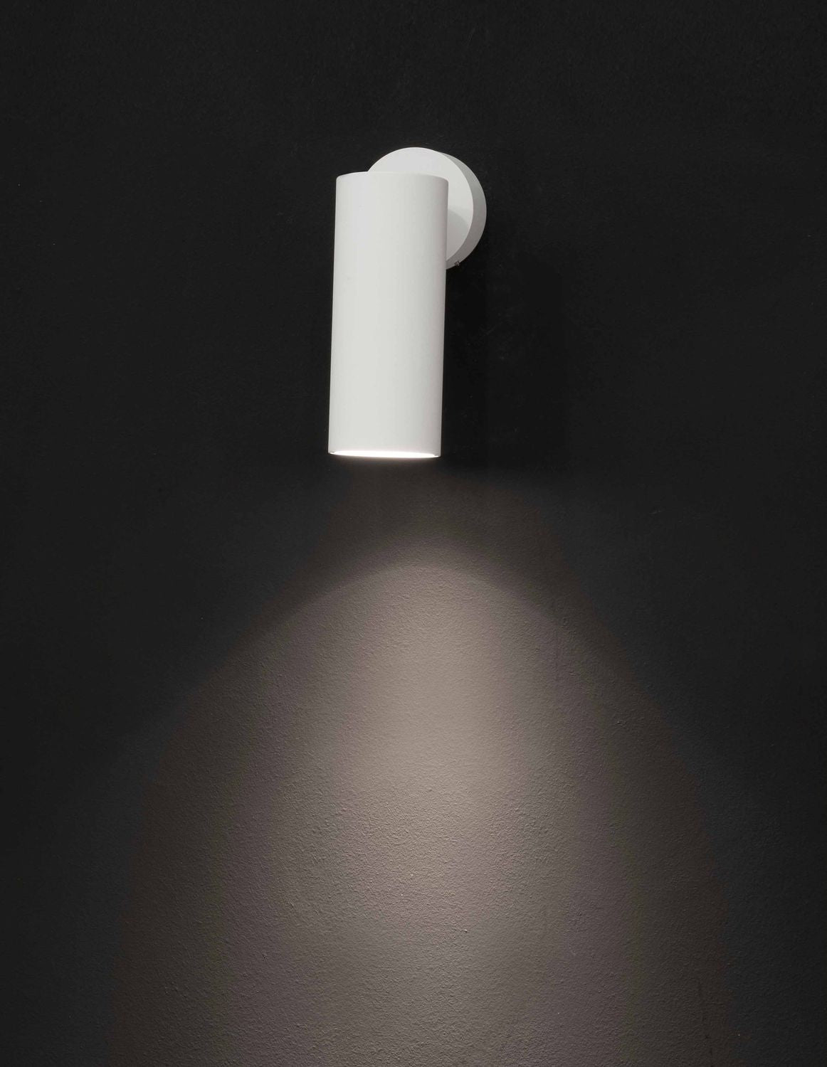 Applique murale blanche cylindrique allumée, projetant un halo lumineux sur un mur sombre.