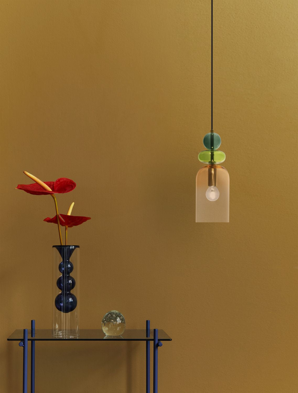 Suspension en verre coloré suspendue devant un mur ocre, près d'un vase et de fleurs