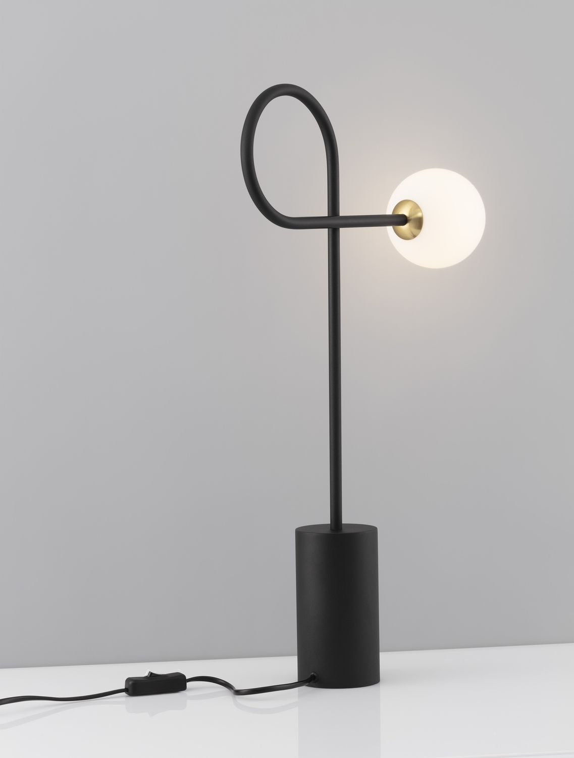 Lampe à poser allumée, base cylindrique noire et globe de verre opalin sur fond gris