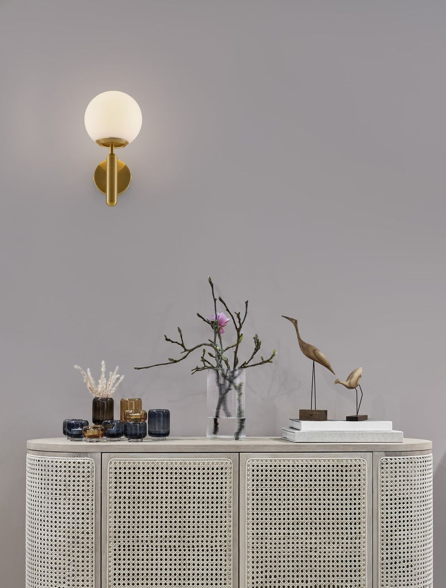 Applique dorée allumée sur mur gris, buffet cannage et objets décoratifs en dessous