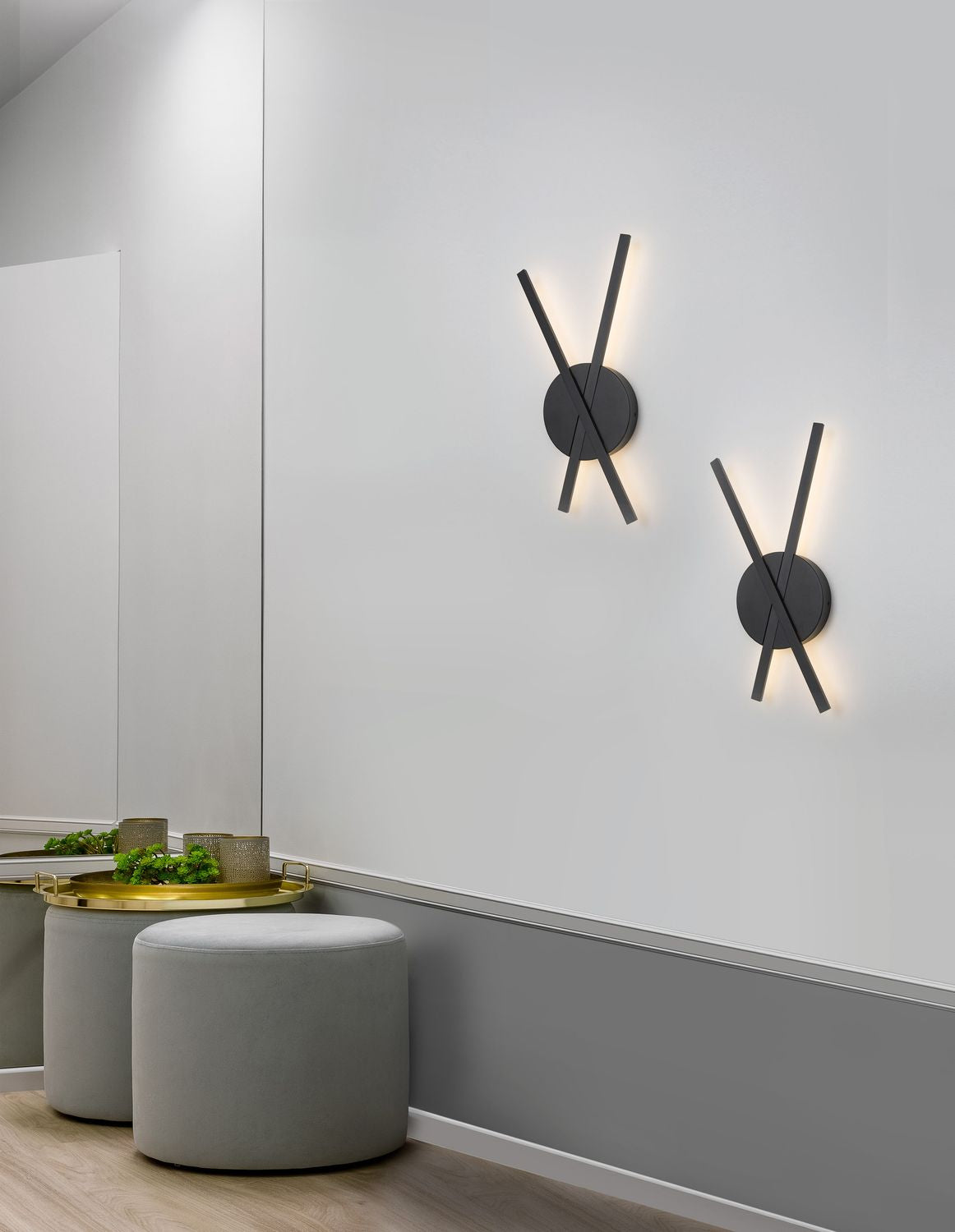 Deux appliques noires allumées sur un mur gris, au-dessus de poufs en velours