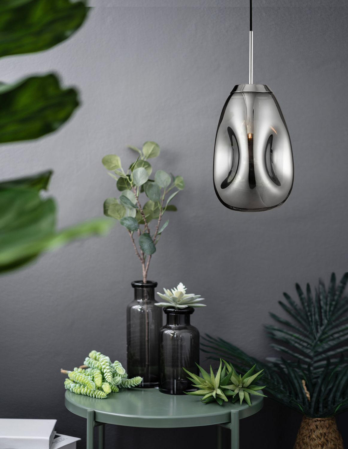 Suspension chromée allumée suspendue dans une ambiance végétale, mur gris et table verte.