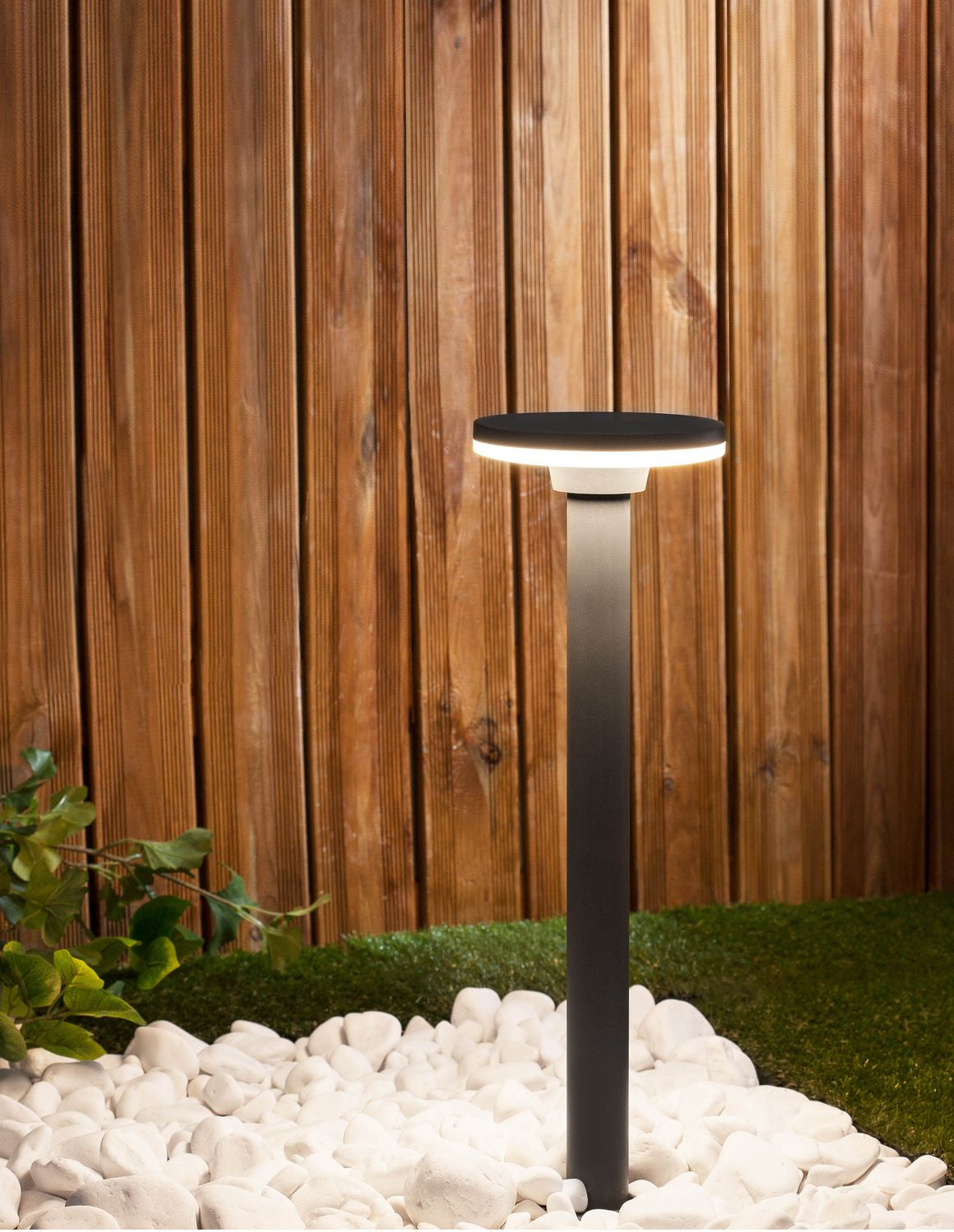 Borne lumineuse grise allumée dans un jardin, gazon, galets blancs et clôture en bois