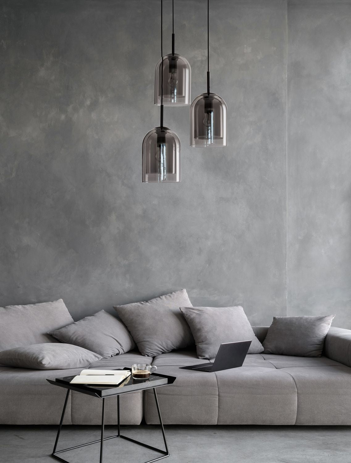 Suspension à trois abat-jours en verre fumé devant un mur gris et un canapé