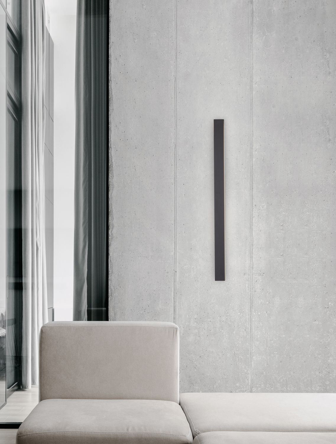 Applique murale verticale noire fixée sur un mur béton, au-dessus d'un canapé beige
