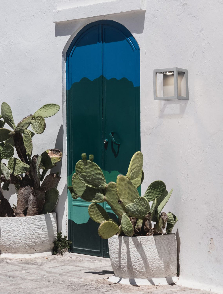 Applique cubique grise fixée sur mur blanc, près d'une porte bleue et de cactus.