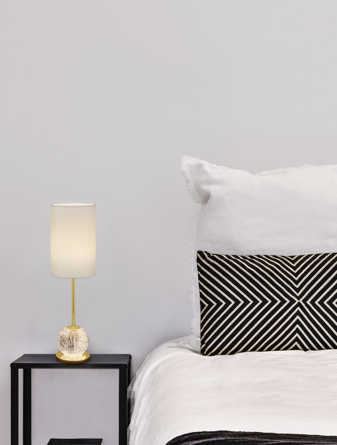 Lampe allumée posée sur une table de chevet noire, ambiance chambre et coussins graphiques