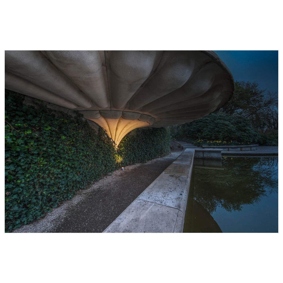 Mise en ambiance nocturne dans un jardin, le luminaire éclaire un mur végétal près d'un bassin.