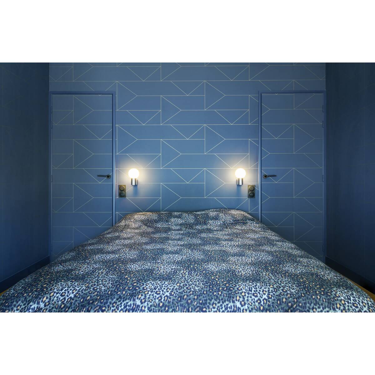 Deux appliques murales éclairent un lit bleu devant un mur géométrique