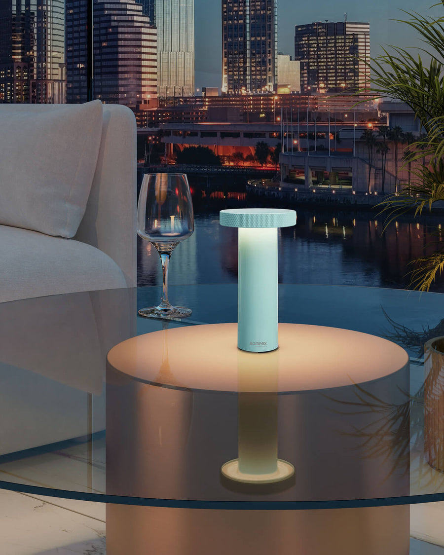 Lampe turquoise allumée posée sur table en verre, vue nocturne urbaine avec canapé et vin.