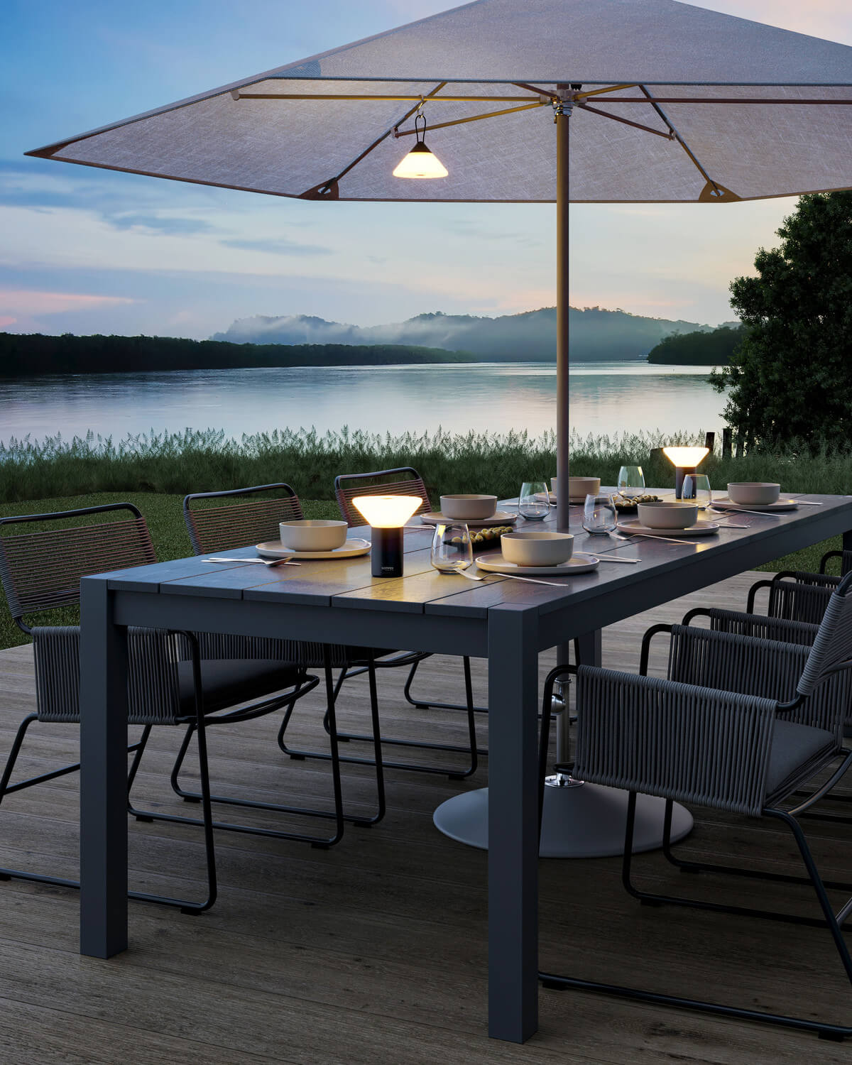 Table extérieure dressée sous parasol au crépuscule, deux lampes posées éclairent le repas.
