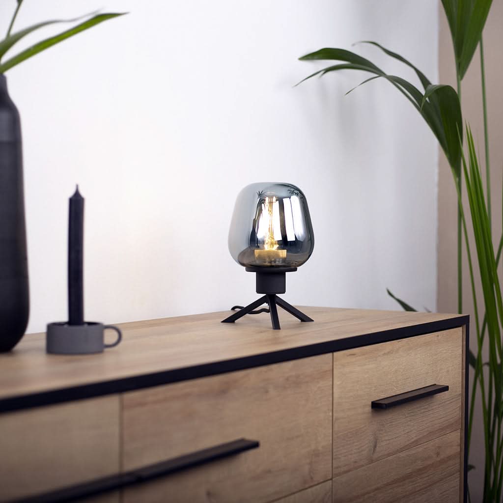 Lampe posée sur commode en bois, allumée, près d'une plante et d'un bougeoir noir.