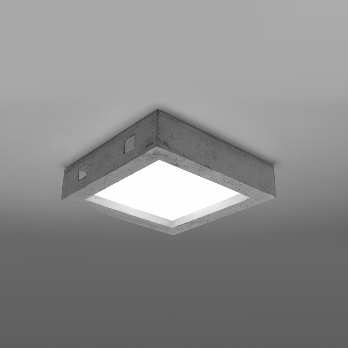 Plafonnier carré en béton allumé au plafond, diffuseur blanc lumineux, ambiance grise.