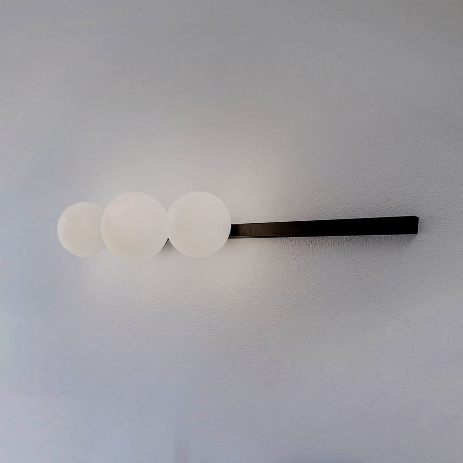 Applique murale avec trois globes en verre opalin blanc sur un long support noir.