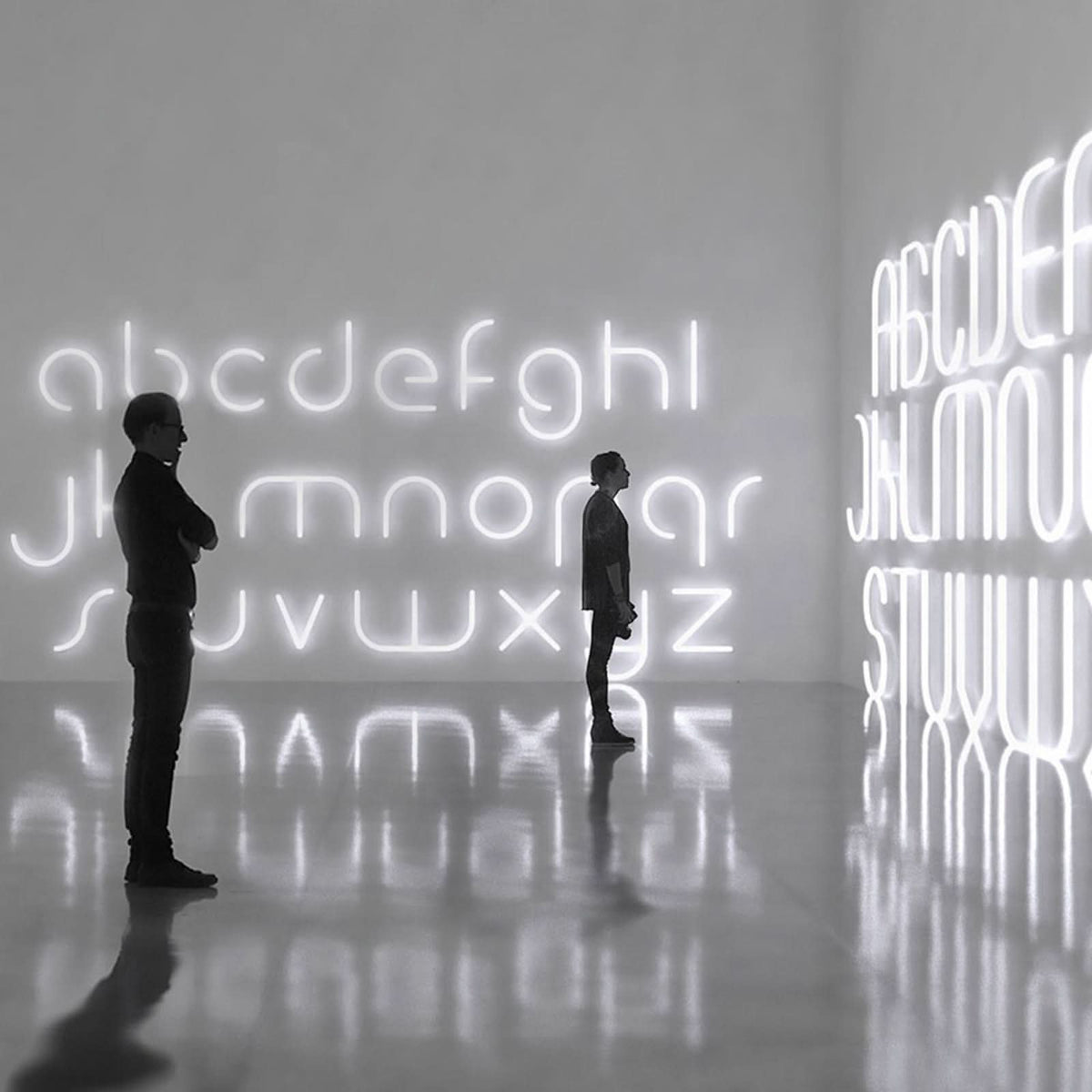 Mise en ambiance dans une galerie sombre avec deux silhouettes face à des lettres lumineuses.