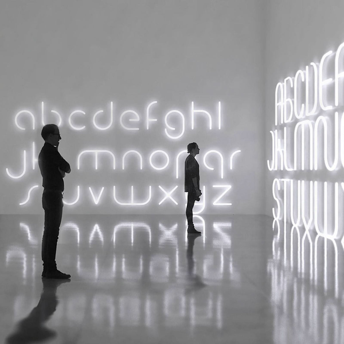 Deux silhouettes observent des lettres lumineuses géantes dans une galerie aux reflets.