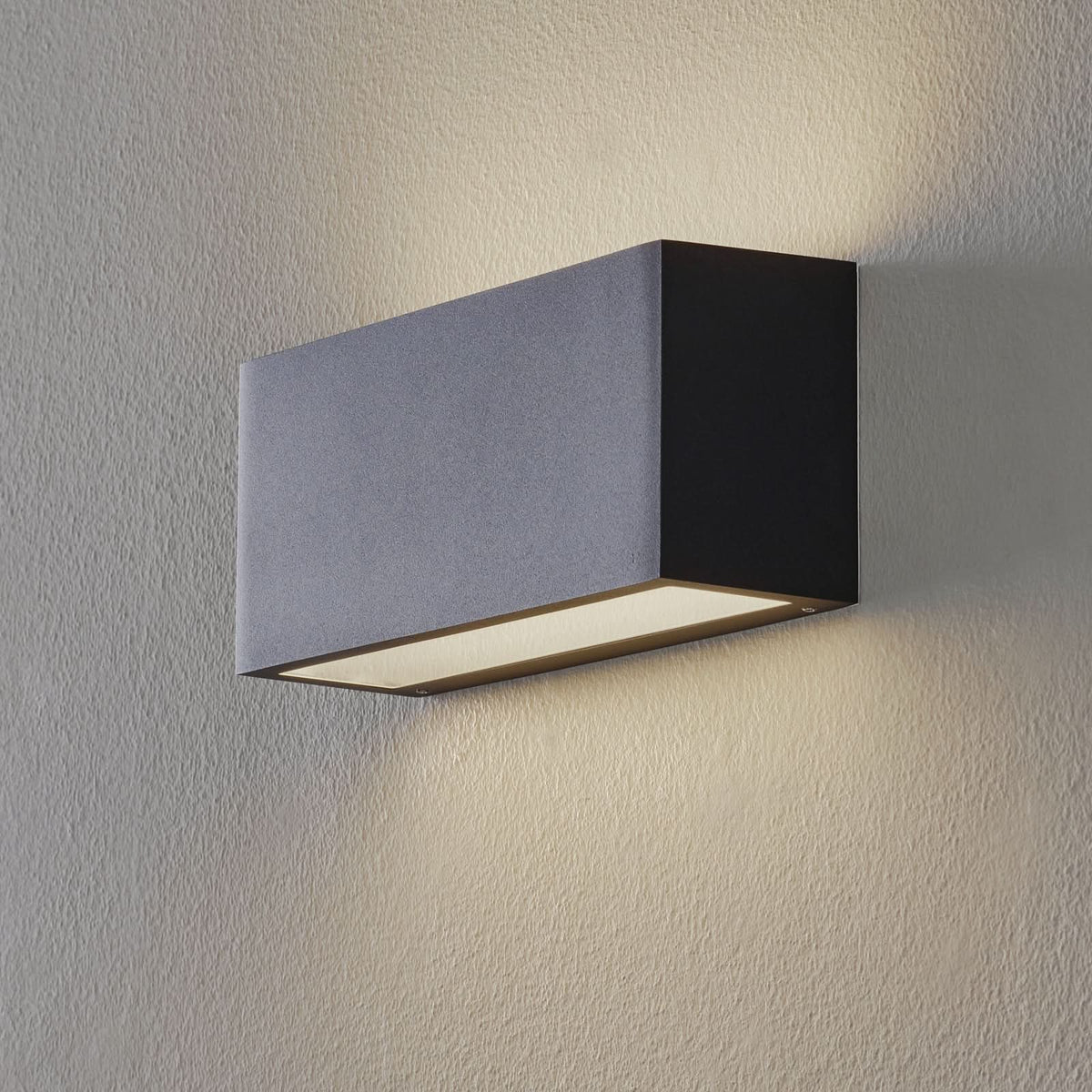 Applique murale rectangulaire noire allumée sur un mur texturé, lumière orientée vers le bas.