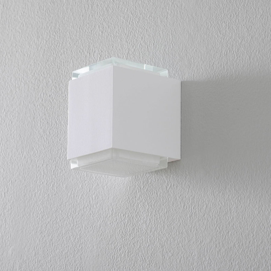 Vue de face du luminaire mural blanc éteint, fixé sur un mur gris clair texturé.